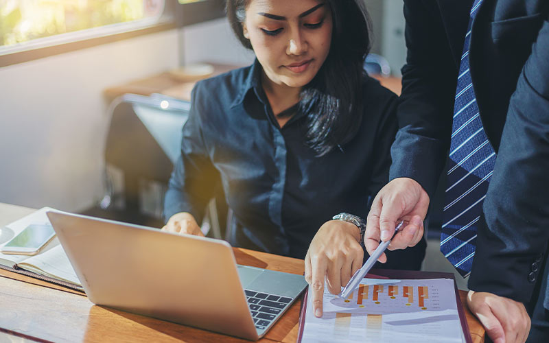 A man and woman in business attire focused on a laptop screen, engaged in professional work.