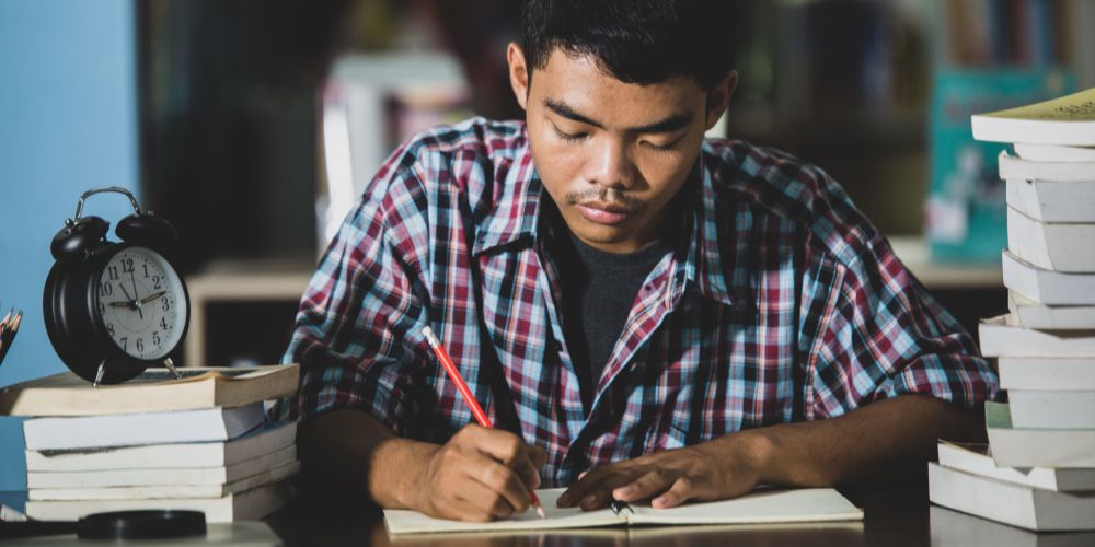 A young man sitting at a desk, focused on writing in a notebook with a pen.