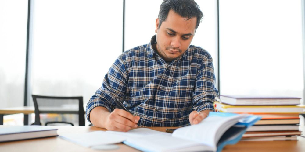 A young man sitting at a desk, focused on writing in a notebook with a pen.